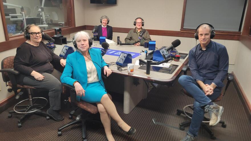 Five people wearing headphones sit at a table in a radio talk studio: a woman front left has short brown hair and is wearing glasses, a black top and grey pants; a woman front right has short grey hair and is wearing a turquoise blazer, turquoise skirt, white shirt and black flats; a woman back left has short grey hair and is wearing a black blazer, purple shirt and white clerical collar; a man back right has short brown hair and is wearing a green fleece over a green button-down shirt; a man at center has short dark hair and is wearing a blue button-down shirt, jeans and sneakers.