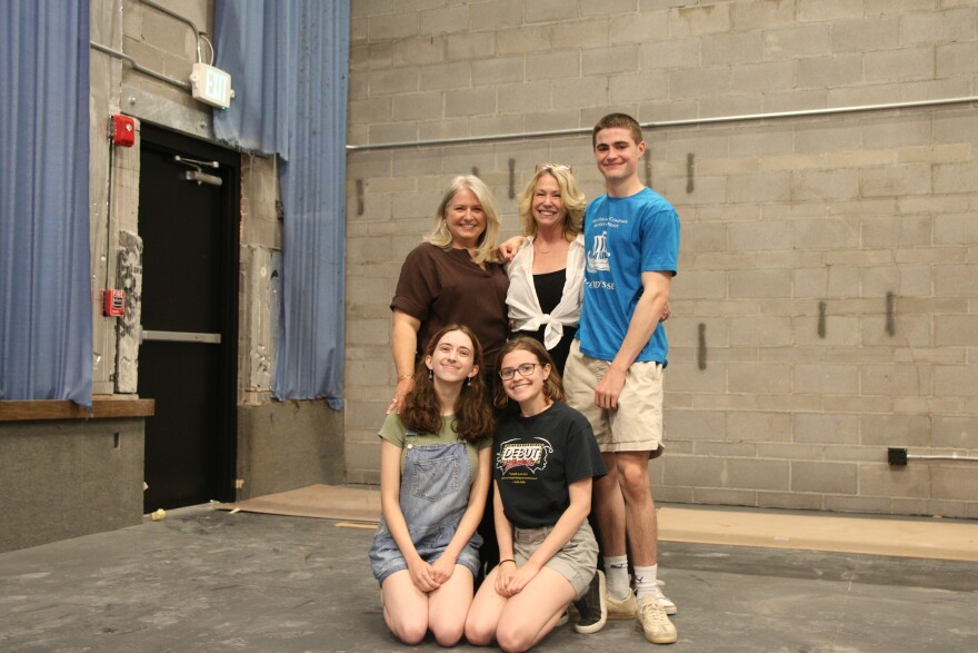 Five people - two adults and three minors - stand and pose for a photo on a stage. Behind them is a partial blue curtain and a concrete wall.