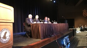 Three men and one woman sit at a table on a stage. 