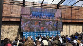 A crowd of people stand in front of a table of eight other people who are eating burritos piled up in front of them.