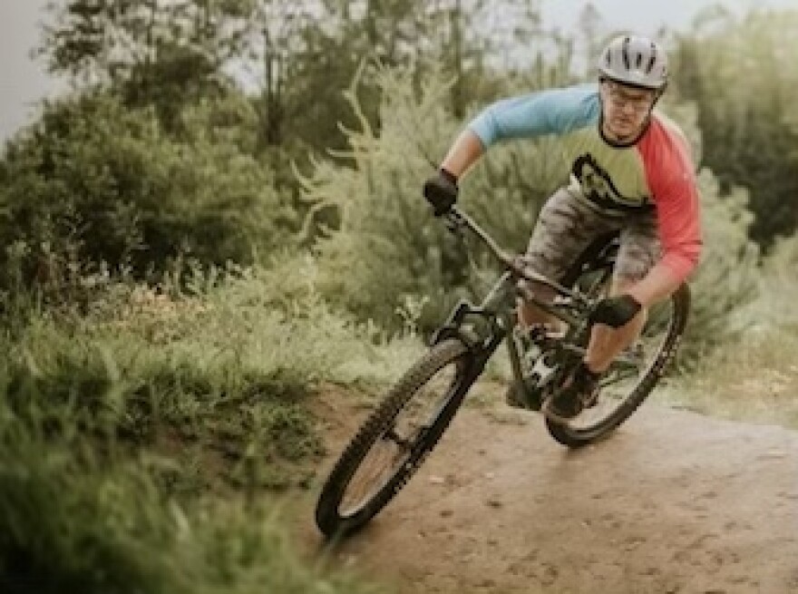 A person wearing a bike helmet, bike gloves, camo-print shorts and a red, white and blue long-sleeved tee shirt rides a mountain bike on a dirt trail in the woods.