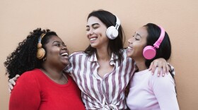 three girls stand in a line with headphones on, laughing