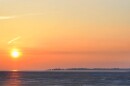 A vivid sunset over a lake with an orange, slightly clouded sky. The sun reflects on the lake. Across the lake, a forest is visible on a far-off shore. 