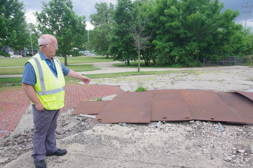 A man in a blue button up, dark grey pants, and a bright chartreuse yellow safety vest stands at the left of the image. He's gesturing towards a section of the road covered by rusted metal covers, surrounded by chucks of asphalt and rocks.