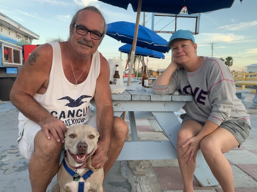 a ban in a white tank top and women in a blue hat sit at a light blue picnic table, A blue beach umbrella is in the background. A smiling dog is in the foreground.