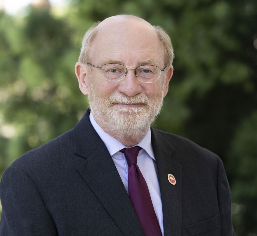 A man in a suit and dark purple tie looks into the camera. He is wearing glasses, a pin is on his left lapel. 