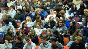 A large group of teenage students in auditorium seats chatting with one another.