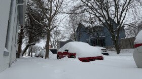Sidewalks, the road and several cars parked in the street are nearly covered in snow.