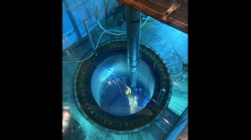 The image is shot from above a pool of blue water. A circular hole sits inside, just below other parts of the submerged metal. A silver metal rod can be seen above the water holding a smaller metal rod that is partially submerged. Various pipes and tubes also feed in and out of the pool. 