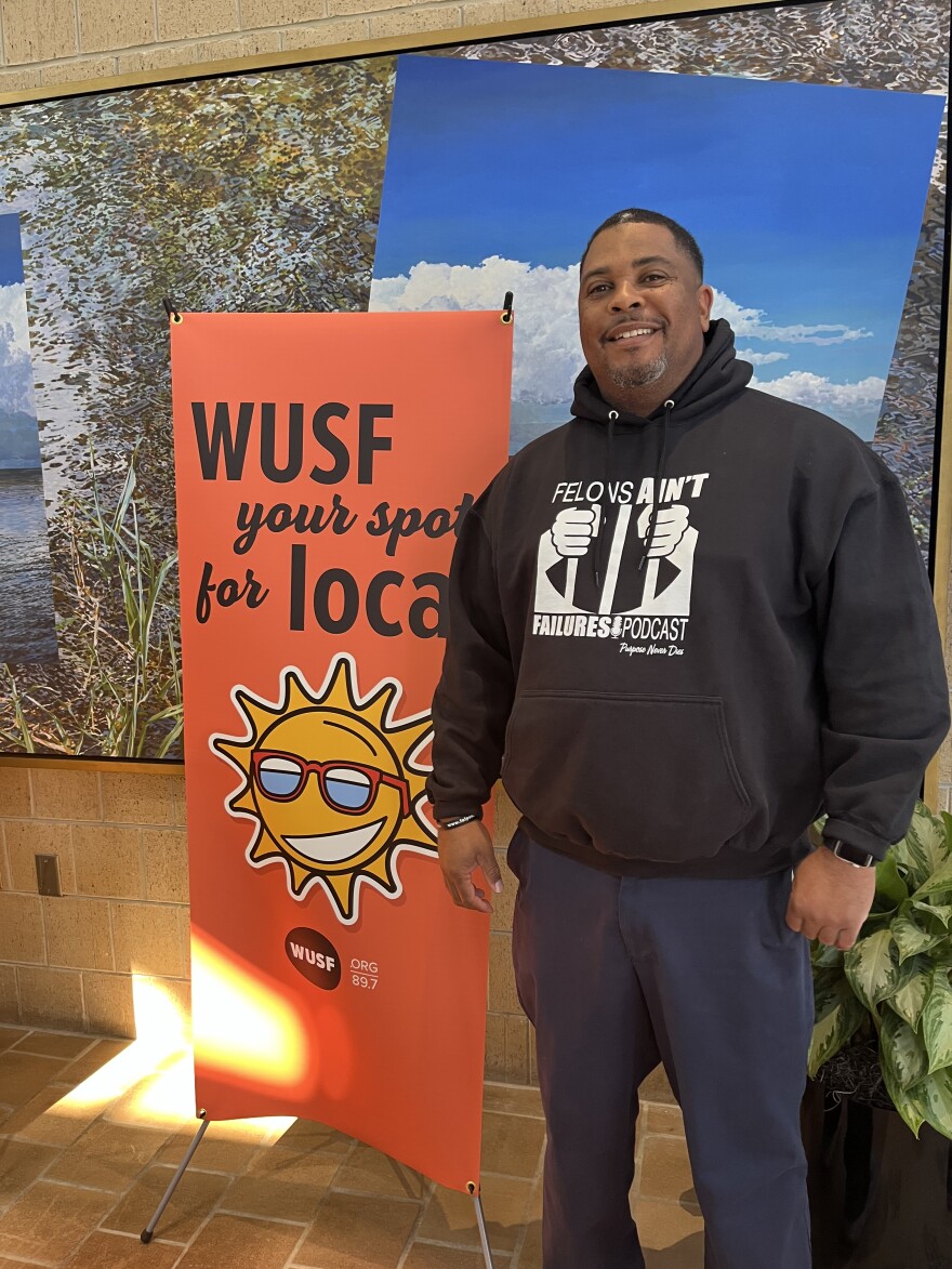 African American man in a sweatshirt that says "Felons Ain't Failures Podcast" stands by WUSF your spot for local pink sign. 