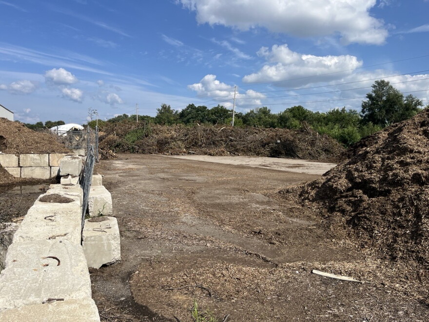 An outdoor compost or mulch facility with large piles of organic material, concrete barriers, dirt ground, and a partly cloudy sky overhead. Trees and power lines are visible in the background.
