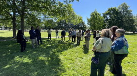 A crowd forms a semicircle standing beneath the shade of a tree in a grassy park.
