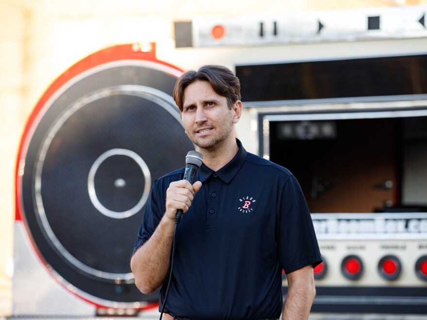 Man in blue polo shirt holding a microphone and standing in front of a large speaker