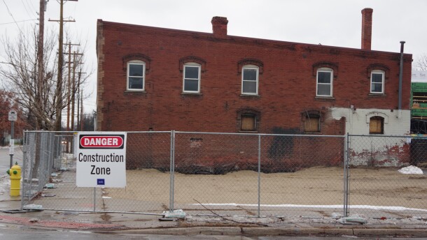 A cold gray chain link fence wraps around a wet patch of sand, a sign declaring the area as a construction zone hangs from the make shift barrier. A dilapidated dark red brick building sits just behind the patch of sand, with light brown ply wood boards a few blocking windows at its base. Small puddles on the adjacent street and gray clouds above denote the rainy weather that occurred that day. 