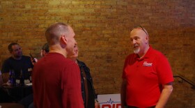 Three people stand in a circle, both men wear red shirts while the woman wears a dark blue denim jacket. A light brown brick wall is illuminated by yellow light behind them. Other attendees can be seen sitting at tables to the left of the image.