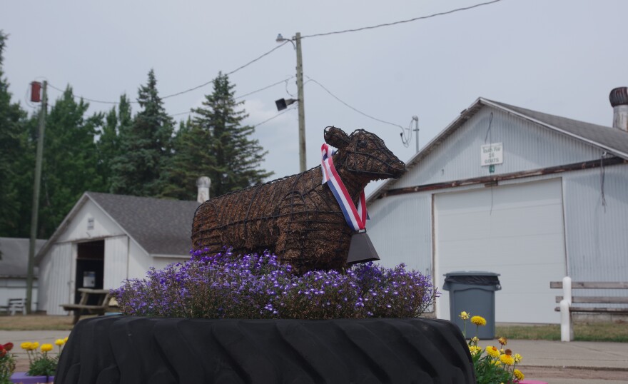 A small statue of a cow sits on top of a large tractor tire. Tiny purple and white flowers surround the statue, having been grown inside the tire. Yellow and red flowers can also be seen in separate pots surrounding the tire. Behind the mock up can be seen two white barns, one on the right of the statue and the other on the left. The left barn's door sits open while the one on the right is closed. The weather is overcast, as clouds fill the sky. 
