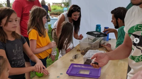 Children gather around a wood-topped table. Two men show the children a variety of insects in their hands and in clear plastic carriers.