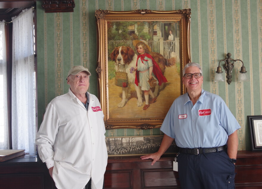 Two men stand in front of an old piece of art work, facing towards the camera. The man on the right is wearing a white lab coat that has the Post logo on left breast area and a baseball cap. The man on the right wears a light blue button-up tucket into dark blue pants. His name, Todd, is written on a tag at the top right side of his shirt, and on the opposite side is another Post logo. The artwork hangs in between them, depicting a young girl in a blue dress and red cape walking with a white and brown St. Bernard dog. 
