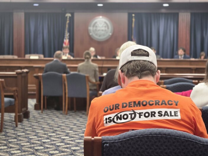 A person in an orange shirt reading "our democracy is not for sale" sits facing away from the camera as a meeting proceeds at the front of a carpeted room.