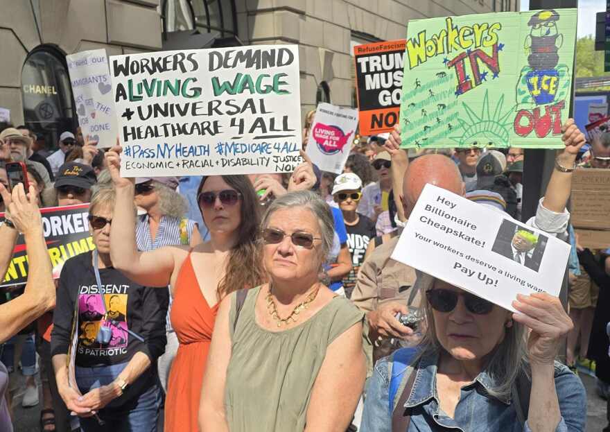 Crowd of people holding signs, workers demand living wage and health care for all and Hey billionaire cheapskate, our workers deserve a living wage. Pay up. 