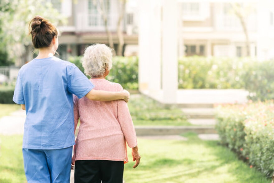 woman in blue scrubs has her arm an older woman with white hair as she escorts her over a grassy green courtyard area, both faces turned away from camera