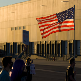 A crowd of people gather outside the ICE warehouse in Salt Lake City. Someone holds up an American flag. 