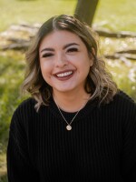 Young Latina female with shoulder length blonde hair, a gold necklace and black sweater, smiling in a grassy area.