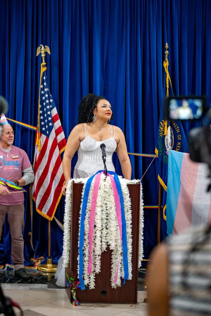 Carma Bell Marshall, wearing a sparkly dress and standing before a podium strewn with white, pink and blue streamers, stands with an American and trans pride flag arrayed behind her at the Kentucky Capitol.