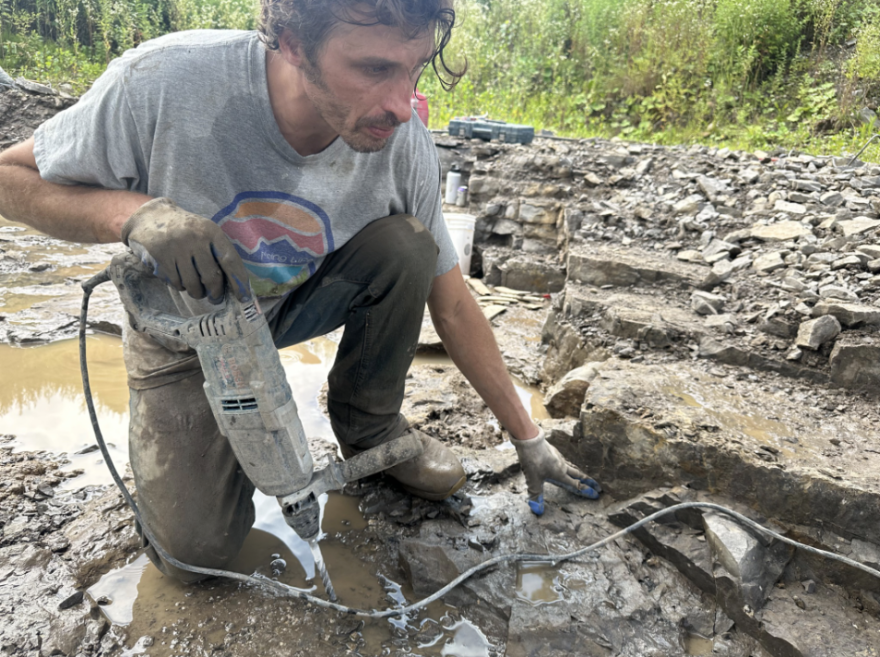 A man in a mud covered t-shirt and canvas pants holds a drill in one hand while hunting for Herkimer Diamonds.