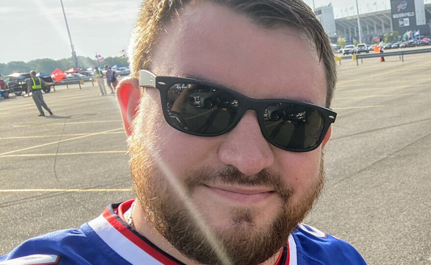 A man with short brown hair and a brown beard stands outside in a parking lot wearing a Buffalo Bills jersey and dark sunglasses.