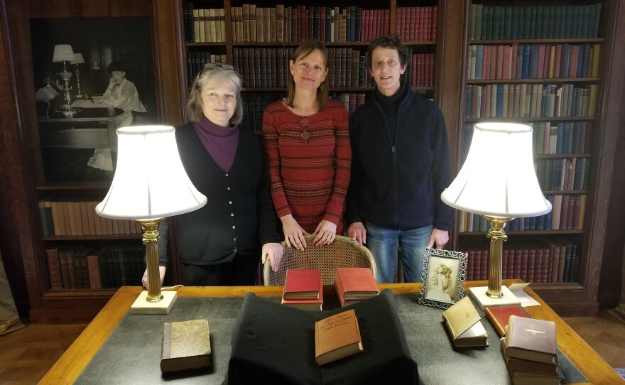 Three women stand behind a desk with a book and two lamps on it in front of a wall of books