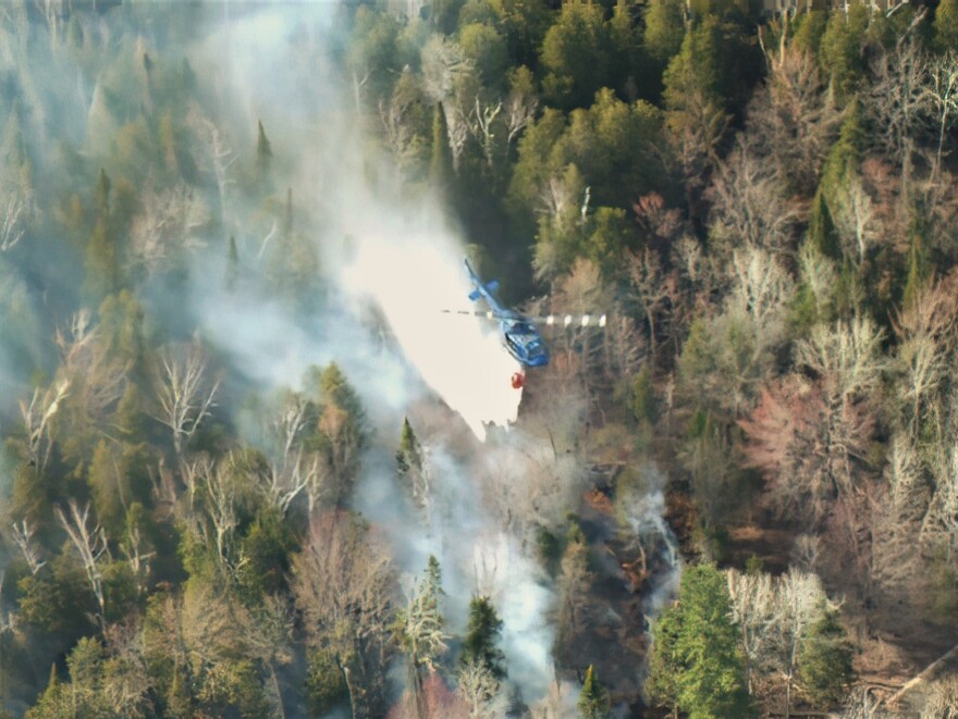 The Michigan State Police UH-1 or "Huey" helicopter drops a 320-gallon load of water over the forest. The bucket is owned by the DNR and the agencies work together to fight wildland fires.