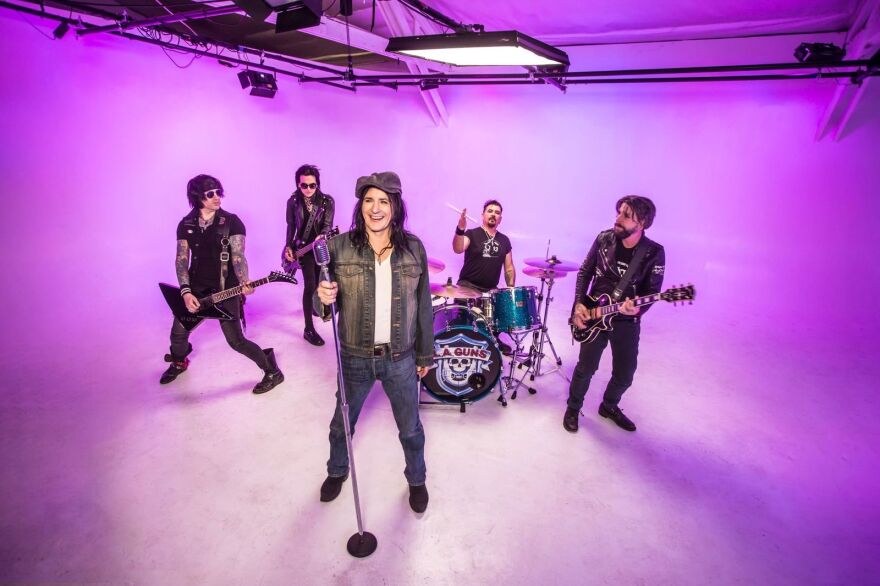 A rock band is performing in a brightly lit studio with a purple background. The lead singer stands at the front with a microphone, while two guitarists, a bassist, and a drummer play behind him.