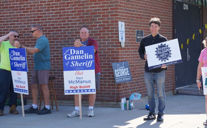 People stand outside a brick school building holding campaign signs