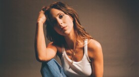 A woman in a white tank top and blue jeans looks at the camera with her elbow resting on her knee and her hand holding her head.