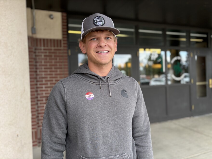 A young man in a gray sweatshirt and baseball hat smiles, wearing an “I voted today” sticker.
