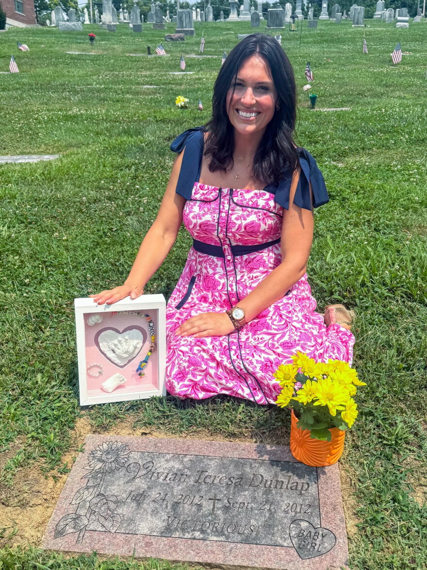 A woman sits behind a headstone holding a shadow box filled with items. a pot of yellow daises sits on the headstone