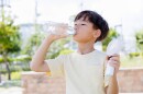 Child drinks water from a plastic bottle while using a portable handheld fan outdoors during a heatwave. 