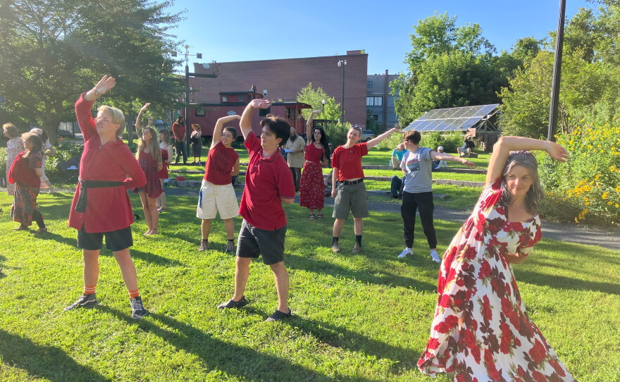  Several people all dressed in red stand in a park with their right arms extended over their heads