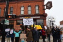 A small group of people stand in front of a crosswalk in downtown Saline. One woman in the center of the image holds a sign that reads “No Secret Deals.”