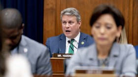 Rep. Bill Huizenga can be seen sitting at a desk with a microphone in front of him and the name placard that says "Mr Huizenga" in front of him. He is speaking in the photo and is wearing a blue suit with a  green tie and a gold lapel pin. He is pictured between two other people sitting at desks. 