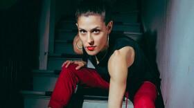 Woman in black boots and a black sleeveless shirt and bright red pants sits on some stairs, leaning forward onto one hand and looking intensely into the camera.