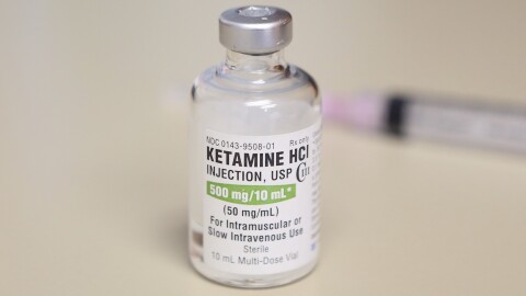 A clear bottle with a label that reads "ketamine" stands on a white table.