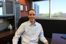 A man in a button down shirt sits at an executive desk in an office and smiles.