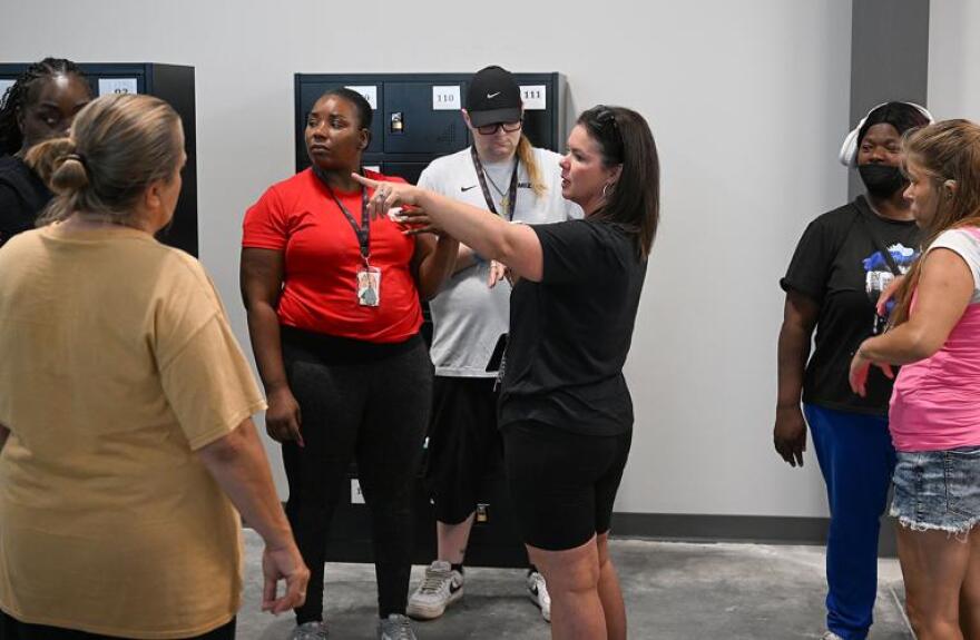 A group of people stand in front of a stand of black lockers as a woman points towards the left.