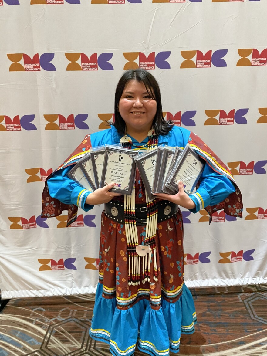 A woman, Jeanette DeDios, poses, showing awards, in front of a backdrop that has a colorful logo and the words, Indiginous Media Conference.