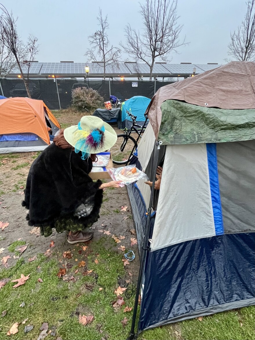 A woman wearing a colorful hat hands a plate of food to a homeless person in a tent. Their hands are visible. 