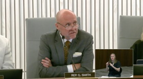 A middle aged bald man with glasses is seated at a table and speaking into a microphone. He's wearing a gray suit jacket and patterned yellow tie. 
