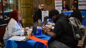 A woman in a light grey sweatshirt stis at a table with a blue tablecloth. A man in a black shirt sits across from her. In the background, two other pairs sit at different tables.