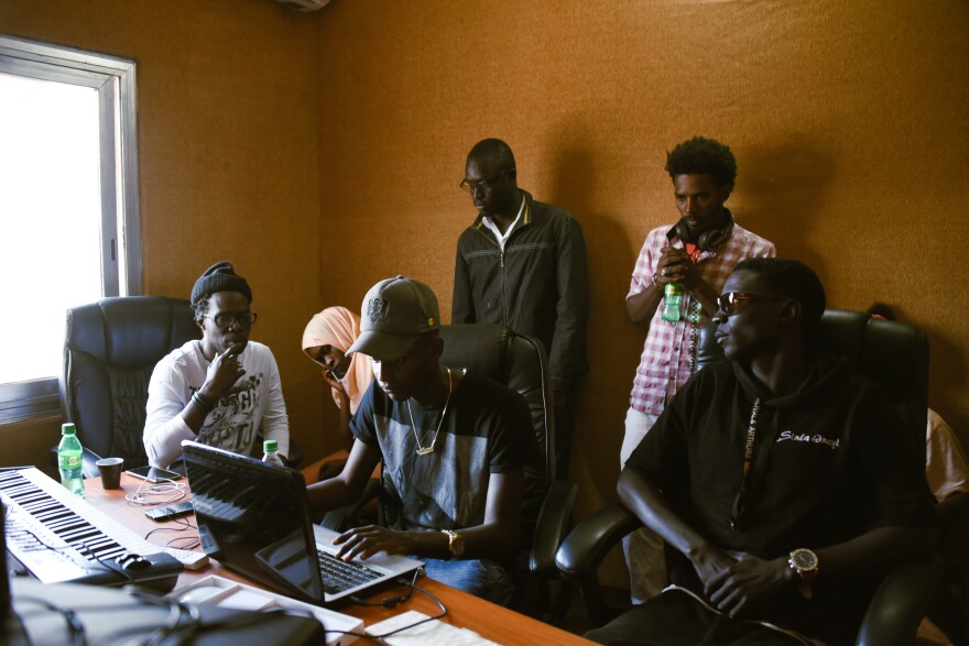 A group of people sit in a recording studio with a laptop and keyboard on the desk in front of them.